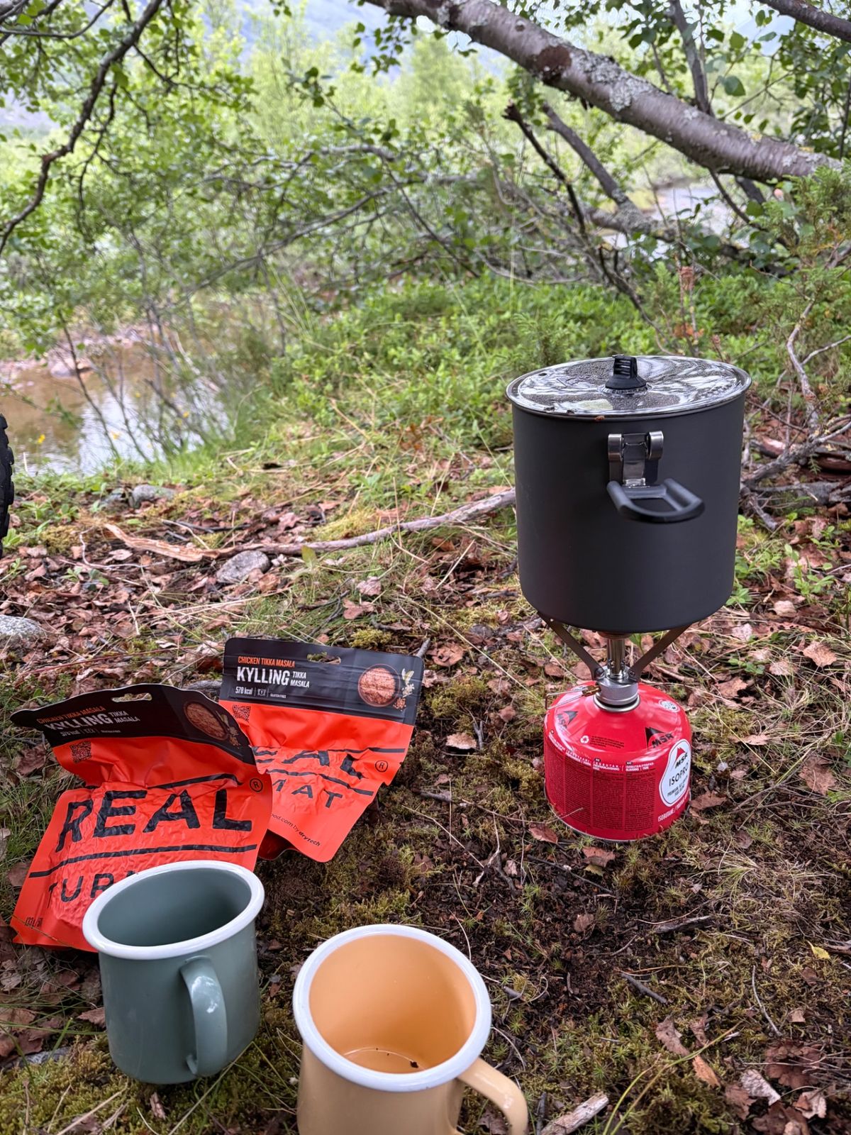 MSR PocketRocket stove with pot and two mugs beside a river