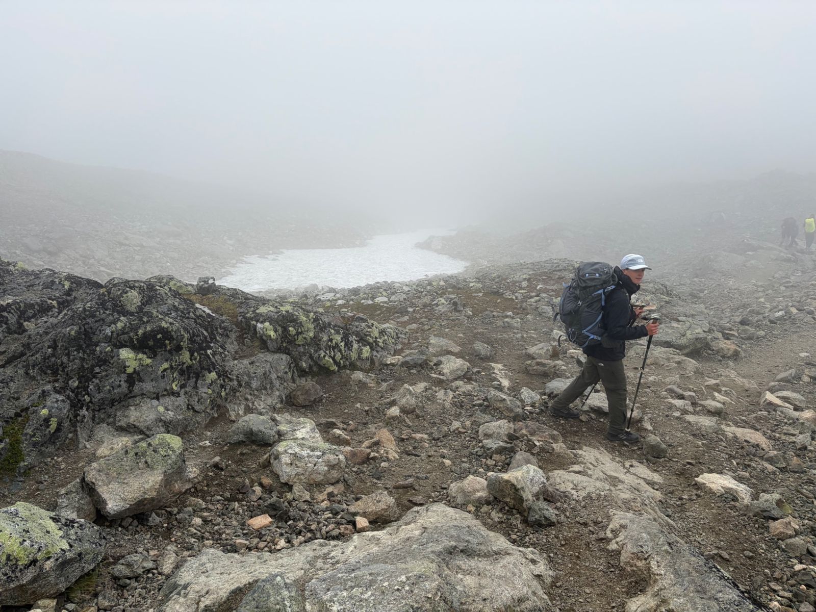 Trekker with pack and poles on a rocky pass in thick fog beside a snowfield