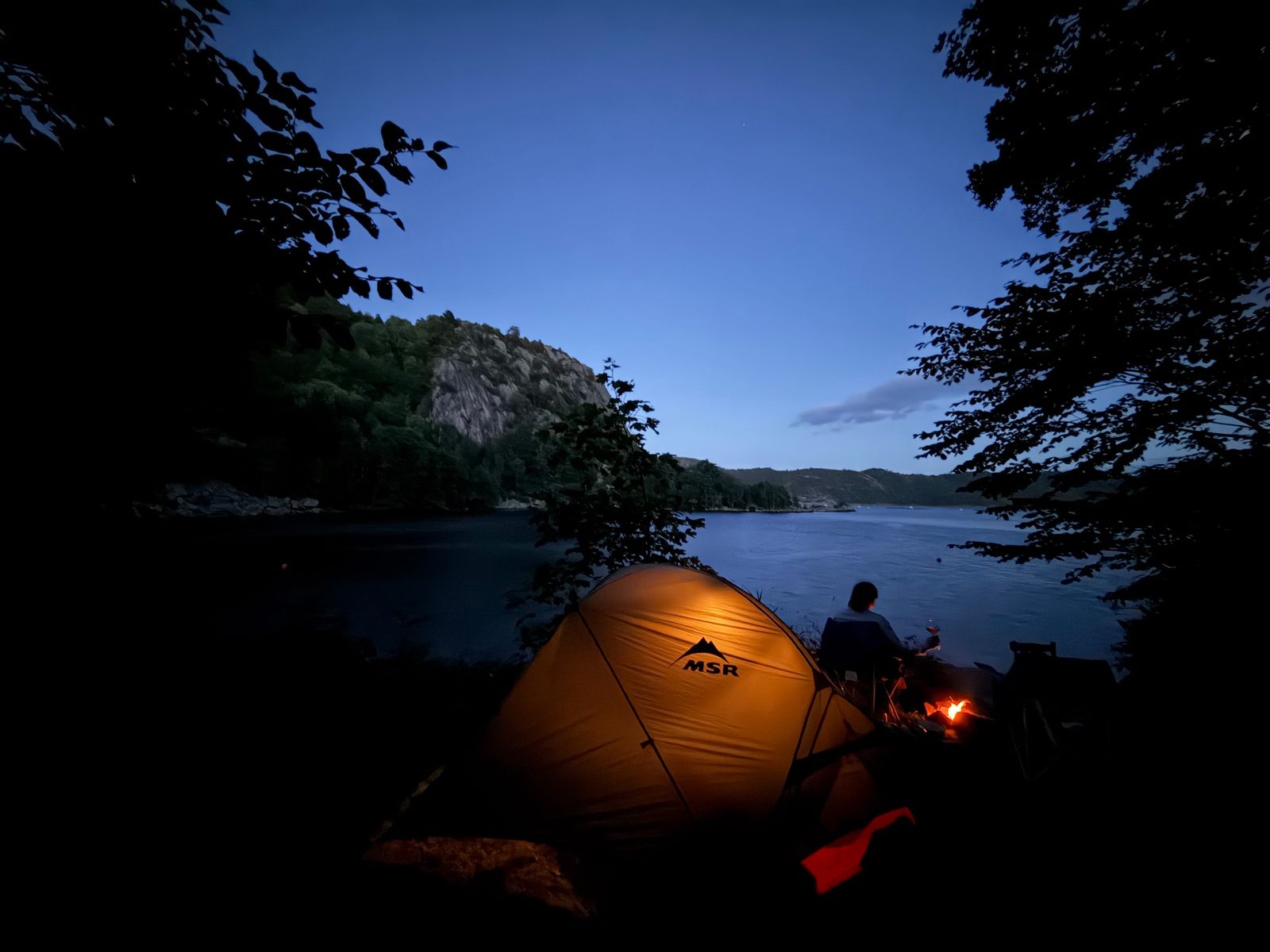 MSR tent glowing at dusk with a small campfire and someone sitting by the fjord